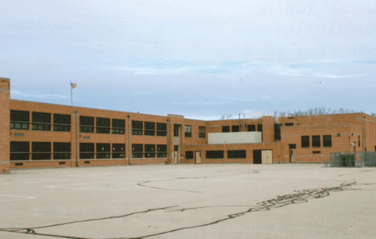 A brick school building, forming a C shape. The majority of the image is dominated by a parking lot with dark ribbons of black tar and faint outlines for basketball or similar. 